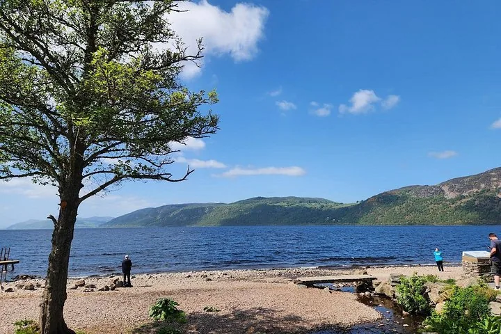 People relaxing on the shore of a loch with rolling hills in the background.