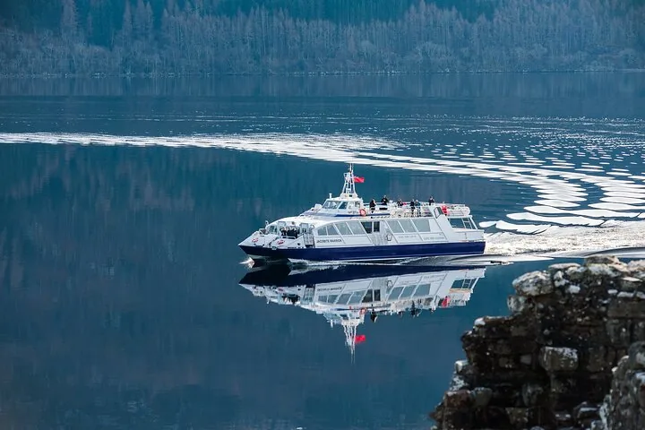 A ferry boat leaves a wake on calm, reflective water near a stony bank.