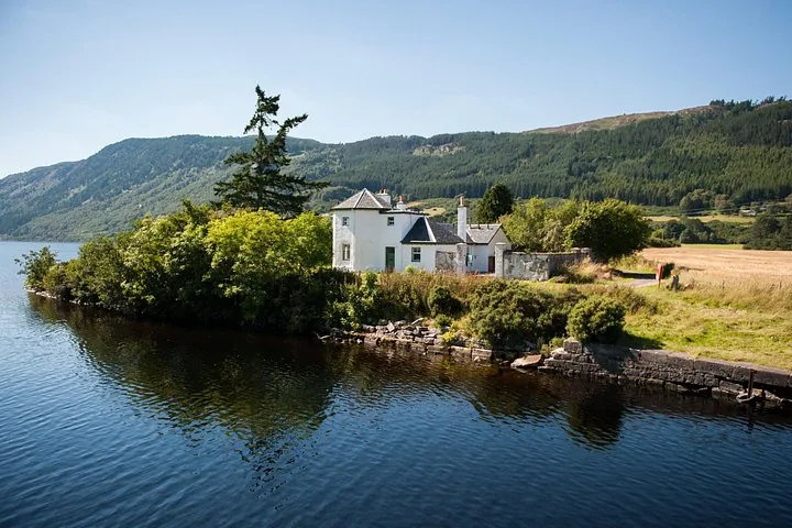 A white house nestled in trees on the edge of a loch.